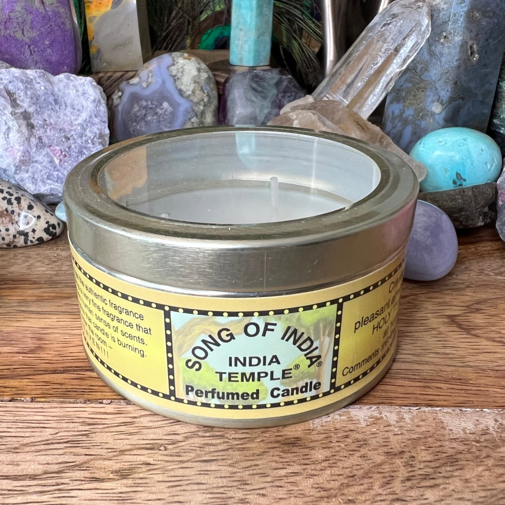 "song of India" candle in a tin with clear lid on wood surface with rocks in background