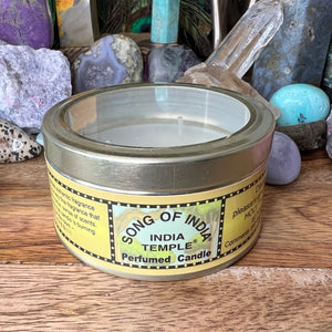 "song of India" candle in a tin with clear lid on wood surface with rocks in background