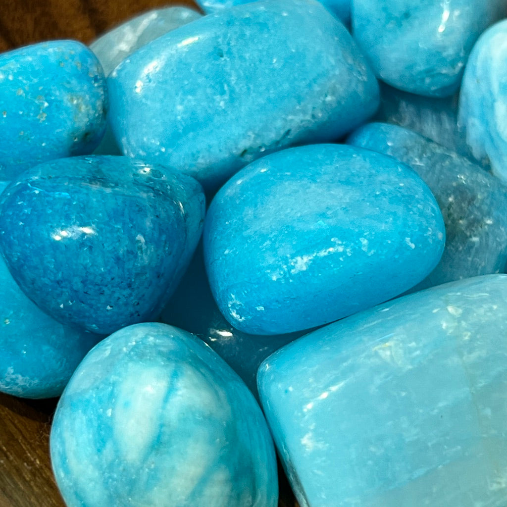 A wooden bowl filled with polished Blue Aragonite sits on a textured surface. Behind the stones, a colorful card reads: "Blue Aragonite - Throat Chakra. Relaxation, eases anxiety, coping with grief and past trauma. Soothes emotional body and aids in vocalizing emotions during emotional transitions.