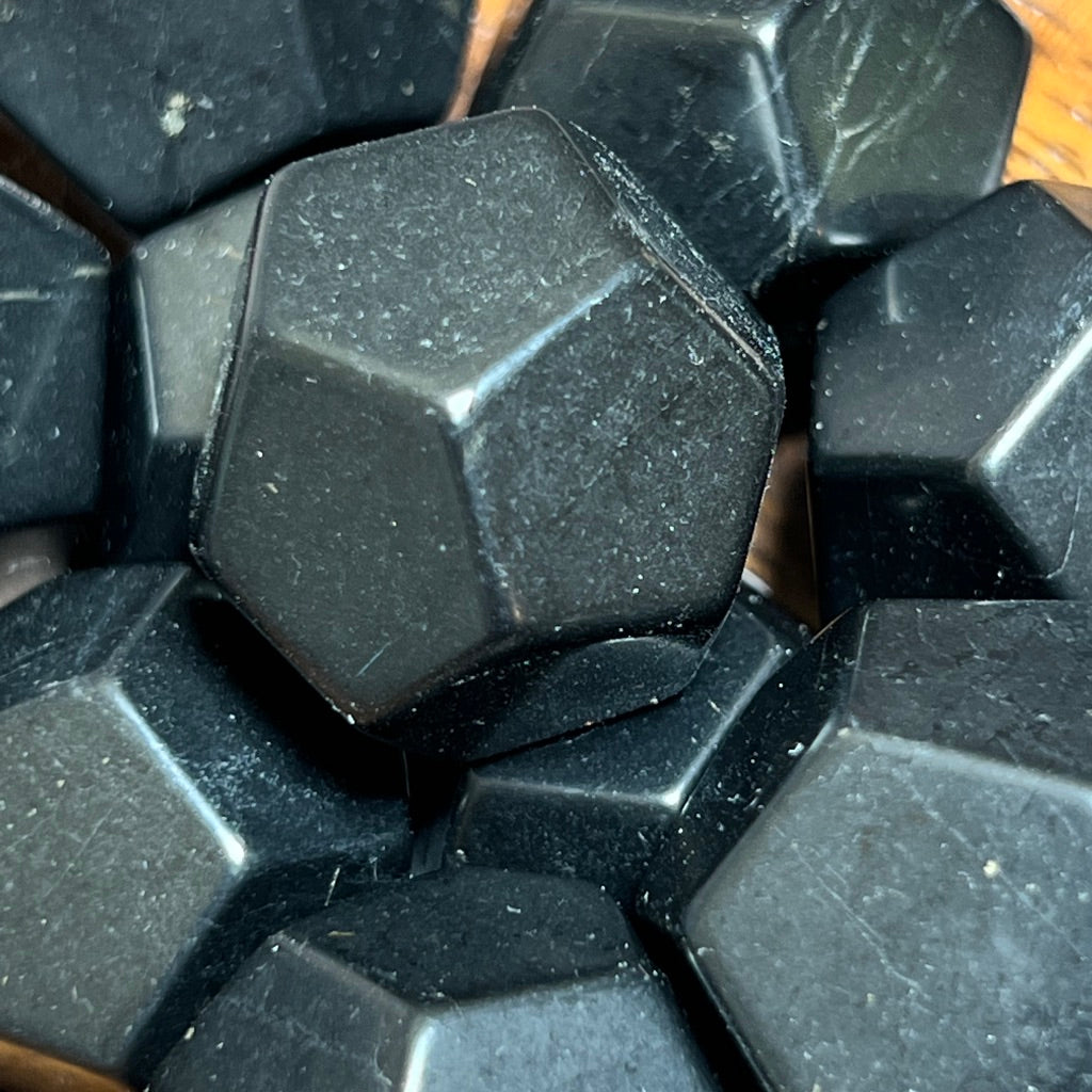 A small wooden bowl holds a collection of Shungite dodecahedrons, reminiscent of Shungite's grounding properties. The bowl rests on a surface with gray, wood-like texture. The dodecahedrons are neatly arranged, showing their faceted surfaces.