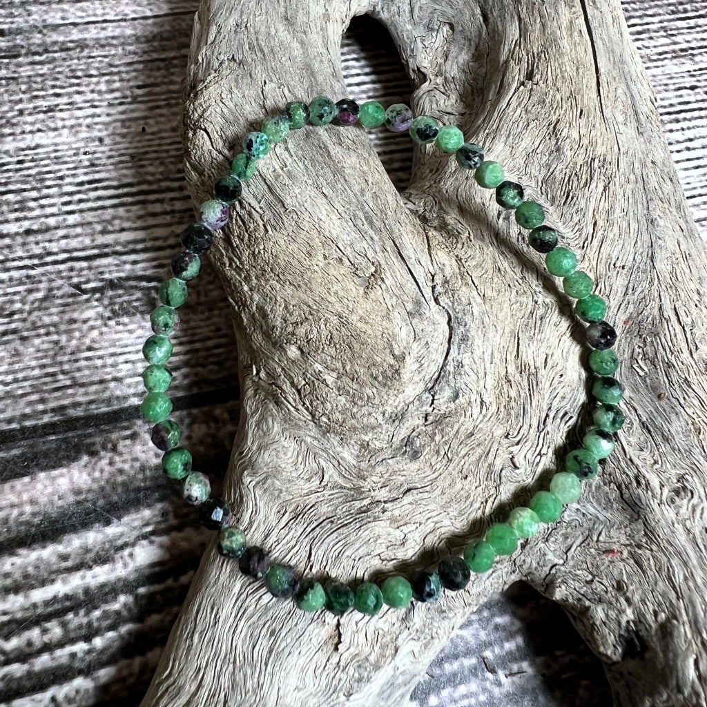 A stretchy bracelet made of ruby zozite faceted beads that are green with black and red specks, displayed on a wooden surface.