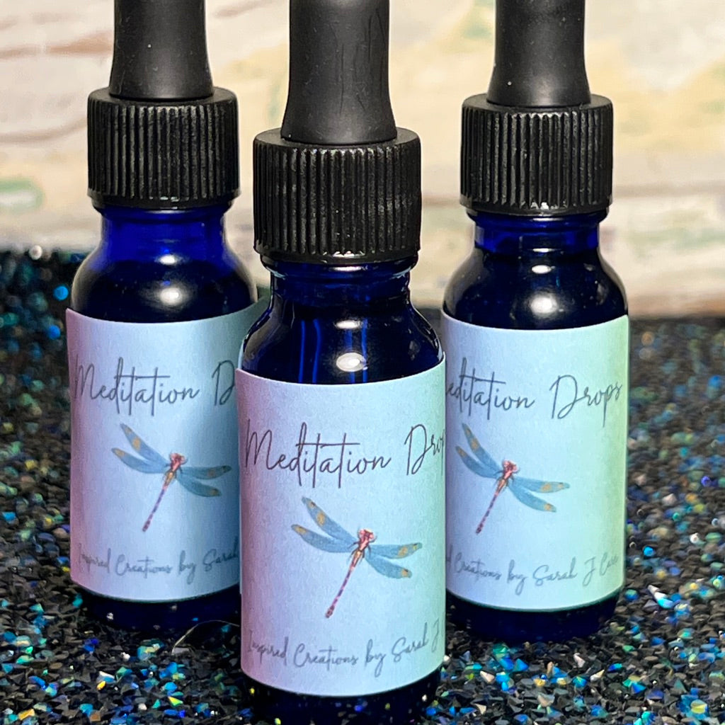 Three small blue glass bottles with black dropper caps labeled "Meditation Drops". The labels feature a dragonfly illustration and read "Handcrafted Creations by Sarah J Ehsani". These bottles, placed on a dark, glittery surface, contain an intuitively crafted blend of high-grade essential oils.
