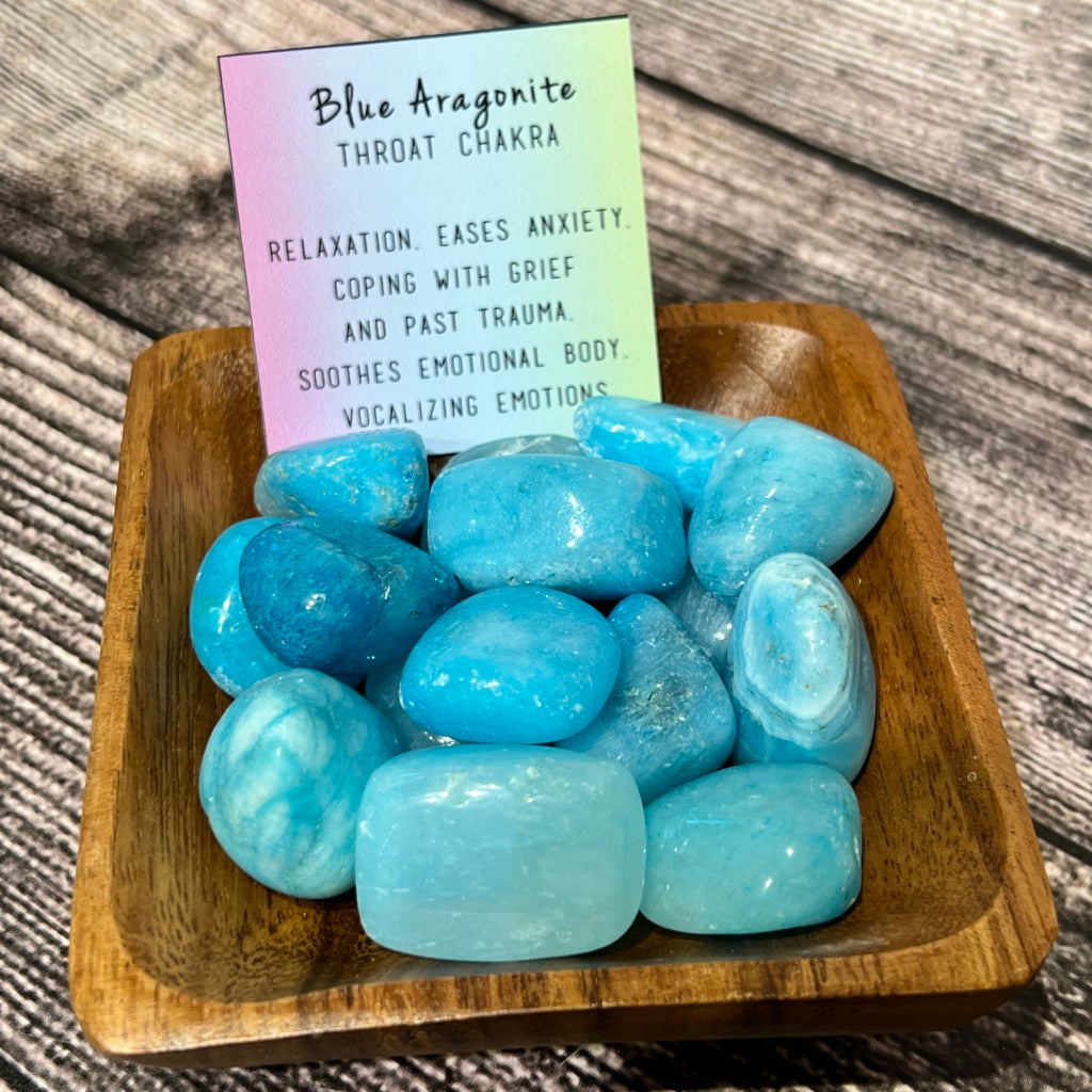 A wooden bowl filled with polished Blue Aragonite sits on a textured surface. Behind the stones, a colorful card reads: "Blue Aragonite - Throat Chakra. Relaxation, eases anxiety, coping with grief and past trauma. Soothes emotional body and aids in vocalizing emotions during emotional transitions.